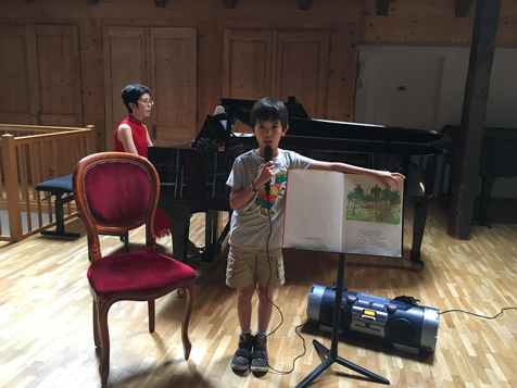 A young narrator holding up an open Babar picture book and speaking into a microphone, with Yuuko Suzuki seated at a grand piano behind him in a wood-panelled hall