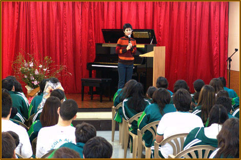 Yuuko Suzuki speaking into a microphone beside an upright piano on a red-curtained stage, addressing rows of seated schoolchildren in uniform