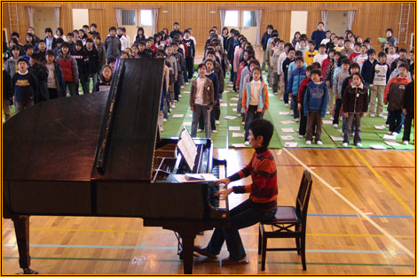 Yuuko Suzuki playing a grand piano in a school hall, surrounded by a large standing audience of children