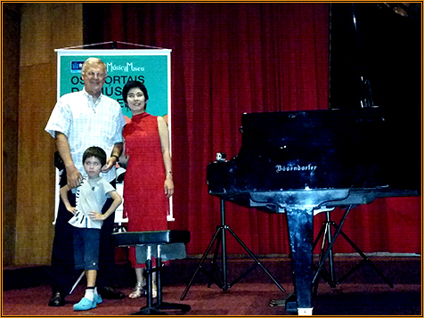 Yuuko Suzuki in a red gown beside a Bösendorfer grand piano, photographed with two others in front of a Música no Museu banner