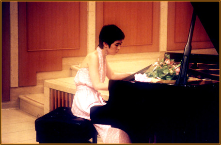 Yuuko Suzuki playing the grand piano in the wood-panelled hall of the Setagaya Art Museum, flowers resting on the instrument