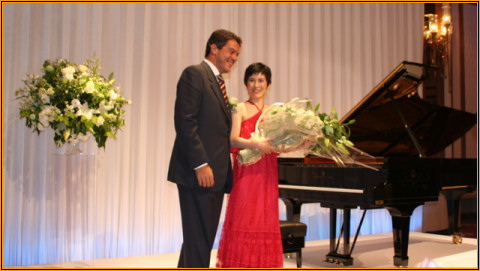 Yuuko Suzuki in a red gown receiving a bouquet on stage beside the grand piano at the Imperial Hotel, Tokyo