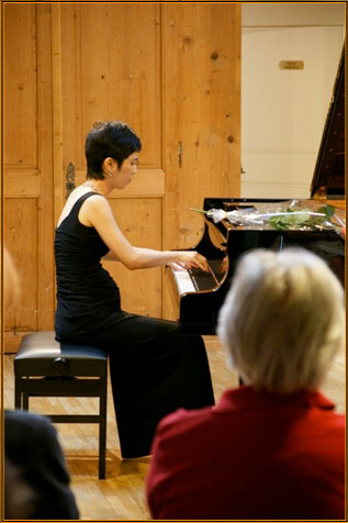 Yuuko Suzuki playing the grand piano in a wood-panelled hall, seen past the heads of the seated audience