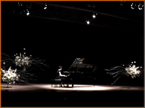 Yuuko Suzuki spotlit at a grand piano on a darkened stage flanked by two large floral arrangements