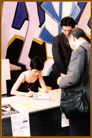 Yuuko Suzuki seated at a signing table with copies of the book, talking with two visitors