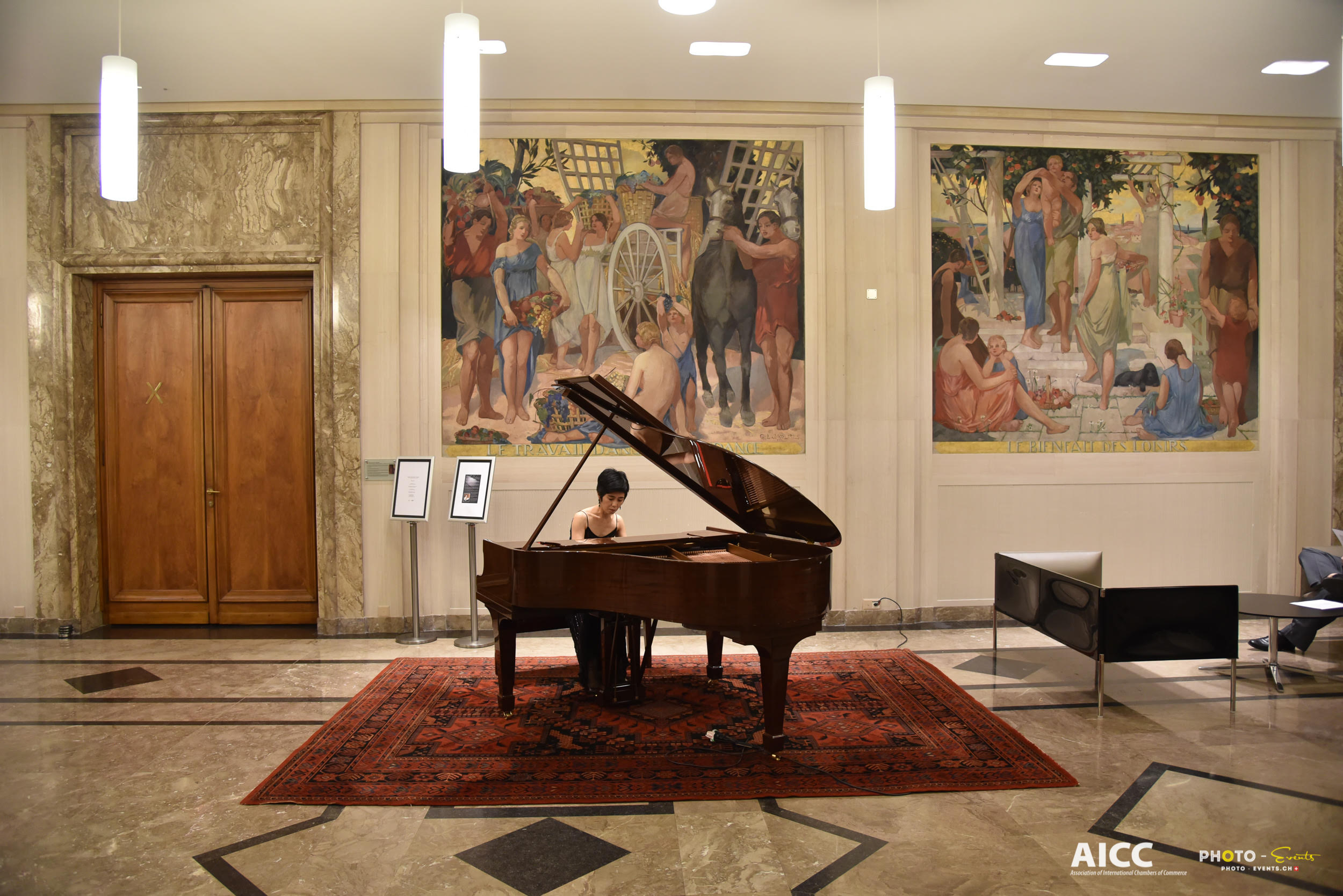 Yuuko Suzuki playing a grand piano on a red carpet in the marble hall of the Centre William Rappard, beneath two large murals